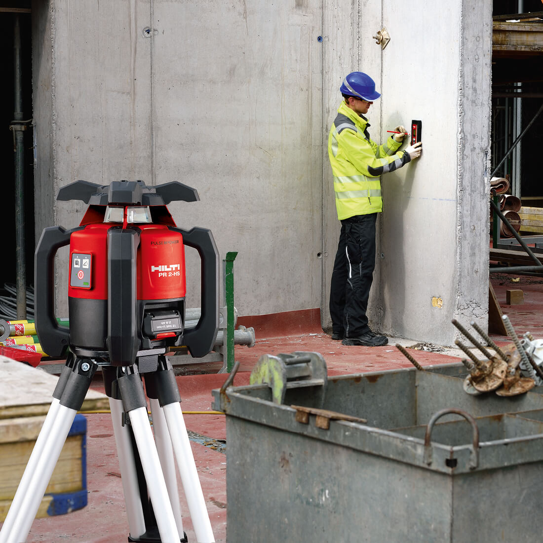 Construction worker in high-visibility jacket and blue hard hat using a laser level to mark measurements on a concrete column; in the foreground a red Hilti rotating laser level on a tripod stands on a construction site floor, product demonstration for KSS Hire.