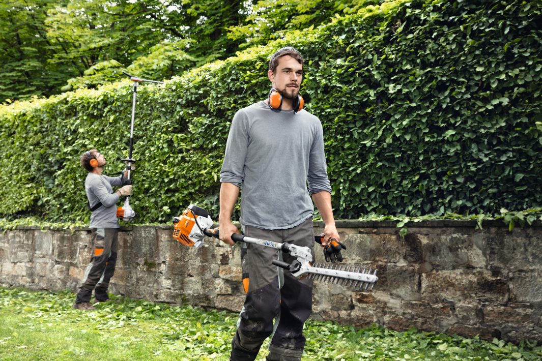 Two workers using long-handled hedge trimmers to trim a tall hedge, with one man in foreground holding the trimmer and another in background