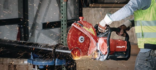 Worker operating a red Hilti disc cutter, cutting a metal pipe with sparks flying, wearing a safety vest